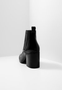 Botines negros de charol con tacón de bloque, con canalé vertical y una pestaña trasera para facilitar el calzado. Textura suave en toda la superficie.