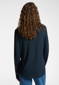 Tummansinisellä pitkähihaisella paidalla on sileä rakenne, kaareva helma ja napitettavat hihansuut. Se on päällä vaaleansinisten farkkujen.