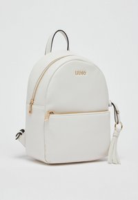 Sac à dos en cuir blanc avec une forme arrondie, des fermetures éclair dorées et une poche avant. Il comporte un pompon décoratif sur le côté. Logo de la marque visible.