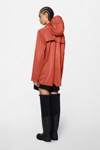 Αδιάβροχο πορτοκαλί αδιάβροχο με κουκούλα, με λεπτομέρεια πτερύγιου στο πίσω μέρος. Φορώντας με μαύρες καουτσούκ μπότες μέχρι το γόνατο.