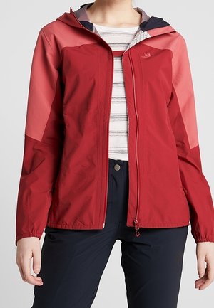 Femme portant une veste à capuche rouge et rose sur une chemise blanche rayée et un pantalon foncé, debout, les mains détendues sur les côtés.