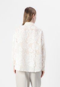 Blusa leggera e trasparente con ricami floreali bianchi su un tessuto chiaro, dotata di colletto e maniche lunghe.