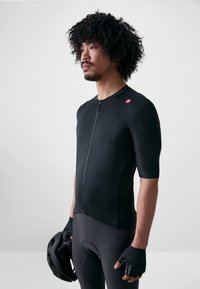 Maglia da ciclismo nera a maniche corte, con zip frontale e tessuto strutturato. Indossata con pantaloni corti da ciclismo neri e guanti, mentre tiene un casco.