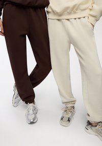 Dwie pary dresowych spodni, jedna w ciemnym brązie, a druga w kremowym, z elastycznymi mankietami i gładką teksturą. Obie noszone z sportowymi trampkami.