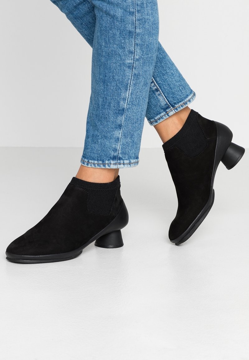 Personne portant un jean bleu et des bottines en daim noir avec un talon bloc arrondi, debout sur un sol blanc lisse.