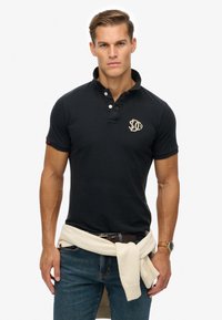 Marineblaues Poloshirt mit Kragen, kurzen Ärmeln und einem gestickten Logo. Kombiniert mit hellblauen Jeans und einem weißen Pullover, der um die Taille gebunden ist.