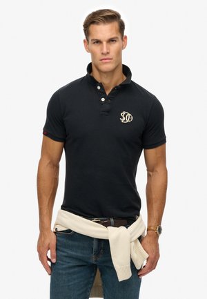 Polo negro de manga corta, con cuello de tres botones, logo bordado en dorado. Usado con un suéter de color claro atado a la cintura.
