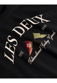 Juoda sportinė striukė su baltais užrašais "LES DEUX" ir "International Sporting Goods", puošti spalvingais išsiuvinėjimais - vėliavėlėmis raudona, žalia ir auksine.