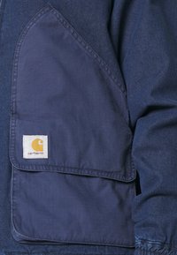 Veste bleu marine en tissu texturé, dotée d'une grande poche plaquée. Logo Carhartt jaune sur une étiquette carrée blanche.