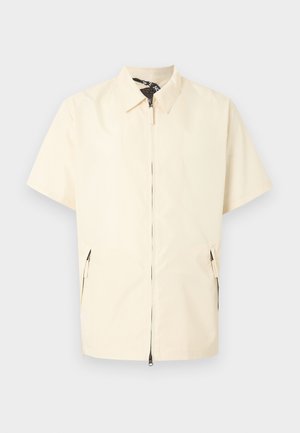 Cremefarbige kurze Jacke mit Kragen, vorderem Reißverschluss und Reißverschlusstaschen an den Seiten vor einem schlichten hellgrauen Hintergrund.