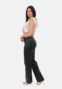 Donna con lunghi capelli scuri, che indossa un top senza maniche in raso beige, pantaloni in ecopelle nera e sandali beige con tacco alto, in piedi di lato.