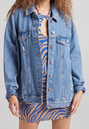 Personne portant une robe à motifs zébrés bleus et beiges sous une veste en jean bleue surdimensionnée boutonnée avec des manches longues.