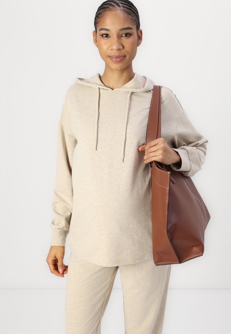 Beige Kapuzenpullover aus weichem Stoff, kombiniert mit passenden Hosen. Braune Ledertasche hat robuste Griffe und sichtbare Nähte.
