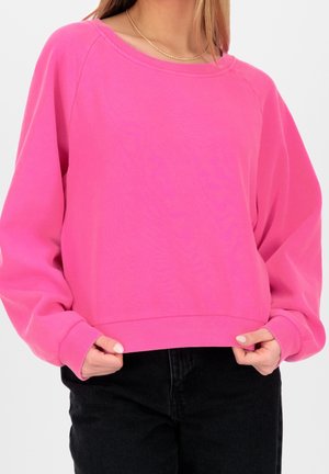 Frau trägt einen knallpinken übergroßen Sweatshirt, hält den Saum, kombiniert mit schwarzen Hosen und einer einfachen goldenen Ketten-Halskette.