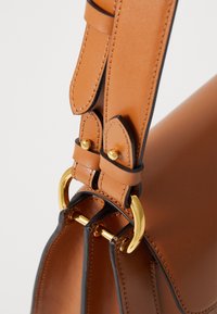 Brown leather handbag strap with gold hardware, smooth texture, double-buckle design, and black accents around the edges.