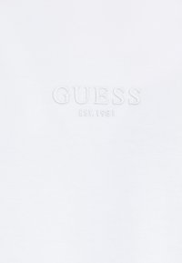 Tessuto bianco con testo in rilievo di colore bianco che legge "GUESS" e testo più piccolo "EST. 1981" sotto.