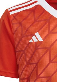 Rotes Sporttrikot mit einem strukturierten geometrischen Muster, weißem Adidas-Logo sowie kontrastierenden weißen Kragen- und Schulterstreifen.