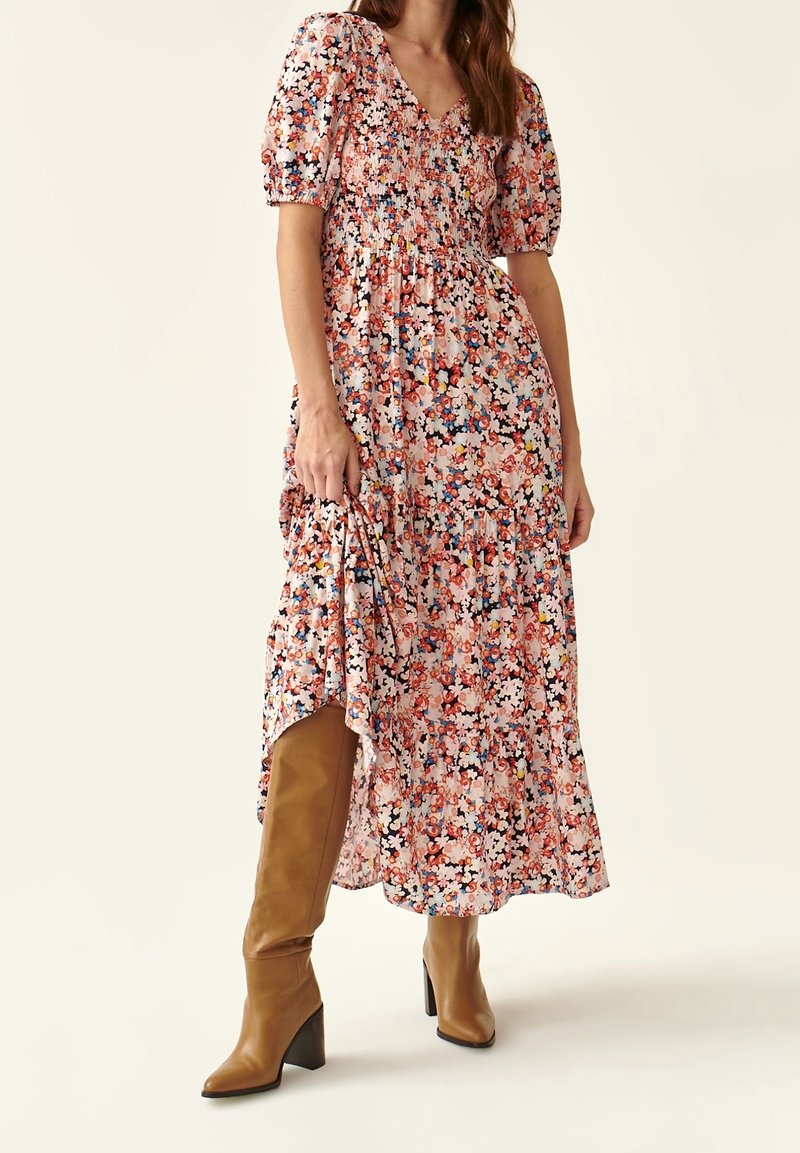 Femme portant une robe midi à manches bouffantes fleurie, associée à des bottes en cuir marron clair montant jusqu'au genou, posant devant un fond uni.