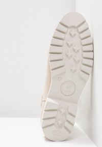 Semelle d'une chaussure beige avec des rainures profondes et des crampons en forme d'étoile, positionnée à la verticale contre un mur d'angle blanc.