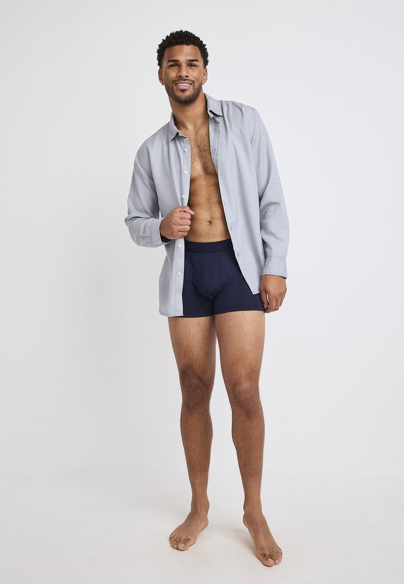 Grå knapp-up skjorta delvis öppen över marinblå kalsonger. Modellen visar korta ärmar och avslappnad passform, står barfota på en ljus bakgrund.