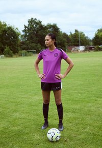 Purple athletic shirt with black accents, black shorts, and knee-high black socks. Standing on grass, next to a patterned soccer ball.