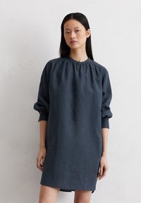 Donkerblauwe linnen jurk met lange mouwen, knoopsluiting aan de voorkant en geplooide halslijn. Zachte textuur met een ontspannen, oversized pasvorm en zijsplitten.