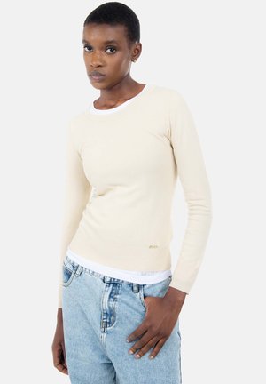 Beige trui met lange mouwen en geribbelde textuur, ronde hals en gouden logodetail, gedragen over een wit overhemd, gecombineerd met lichtblauwe spijkerbroek.