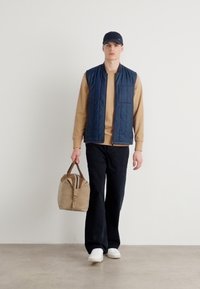 Colete acolchoado azul-marinho sobre um sweatshirt bege, calças pretas, sapatilhas brancas e a segurar uma bolsa castanha clara com um detalhe às riscas.