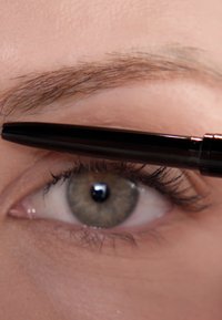 Czarny eyeliner w ołówku jest nanoszony na zbliżenie oka. Oko ma długie rzęsy i jasnobrązową tęczówkę. Tekstura skóry jest gładka.