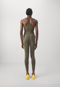 Combinaison de sport vert olive avec un design ajusté, des bretelles croisées dans le dos et une texture lisse. Associée à des baskets jaunes.