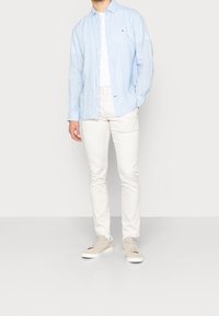 Svetle modrá košile na knoflíky vyrobená z lnu, bílý tričko pod ní, krémové slim-fit kalhoty a béžové tenisky.