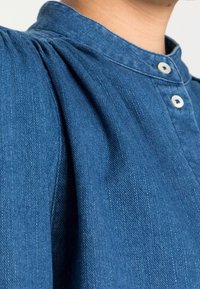 Camicia in denim blu scuro con colletto alto, chiusura a bottoni e una sottile trama con dettagli arricciati sulle spalle.