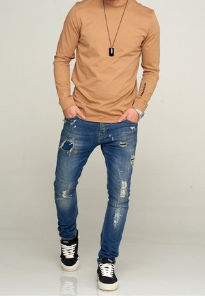 Langärmliges beige Hemd mit Druckdetails an den Ärmeln, kombiniert mit abgewetzten blauen Jeans und schwarzen Sneakern mit weißen Sohlen.