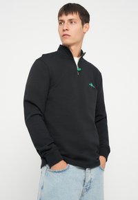 Crna zip-up dukserica s visokim ovratnikom, zeleni logotip na prsima i rebrastim manžetama, uparena sa svijetloplavim trapericama.