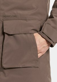 Brun vattentät jacka med en stor framficka och en slät textur. Har sydda detaljer och varumärket "Didriksons" på tyget.