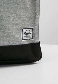 Harmaa kangaskassi, jossa on musta pohja ja jonka sivussa on Herschel-logotägi. Tekstuuri näyttää kudotulta, mikä tarjoaa rakenteellisen muotoilun.