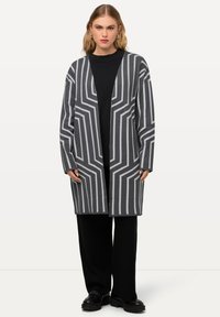 Grauer langer Cardigan mit weißen geometrischen Mustern, offener Vorderseite und lockerer Passform, kombiniert mit schwarzem Oberteil und weiten schwarzen Hosen.