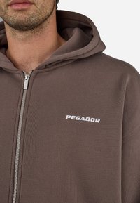 Barna, cipzáros kapucnis pulóver, puha anyagból készült. Ezüst színű cipzárral és fehér "PEGADOR" logóval rendelkezik a mellkason. Rendszeres szabású, kapucnija van.