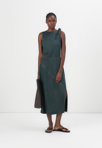 Dark green denim dress with a sleeveless design, asymmetrical hem, and a knot detail on the shoulder, paired with a brown handbag.