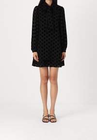 Robe noire à pois réalisée en matière transparente avec des manches longues et un col en nœud. Coupe ample avec un ourlet évasé. Sandales noires à brides.