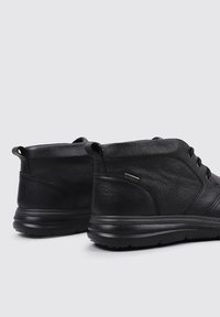 Par de botas de cuero negras hasta el tobillo con cordones en la parte frontal, cuellos acolchados y suelas de goma gruesa sobre un fondo gris claro.