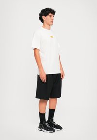 Λευκό βαμβακερό μπλουζάκι με μικρό κίτρινο λογότυπο, συνδυασμένο με μαύρες σορτς. Μαύρα αθλητικά παπούτσια με λευκές λεπτομέρειες και μαύρες κάλτσες μέχρι τον αστράγαλο.