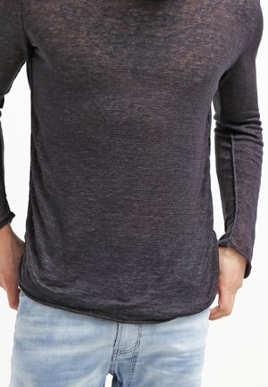 Homme portant une chemise à manches longues semi-transparente gris foncé et un jean bleu clair, les mains reposant près des hanches.