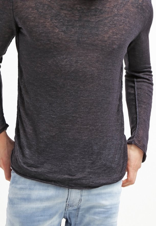 Homme portant une chemise à manches longues semi-transparente gris foncé et un jean bleu clair, les mains reposant près des hanches.