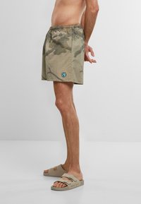 Costume da bagno verde mimetico in khaki con elastico in vita, caratterizzato da una toppa circolare con logo sul lato sinistro. Indossato con sandali beige.