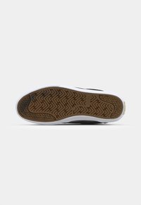 Black sneaker with a white rubber sole. The underside features a woven texture with herringbone patterns. Accents in gold display the brand name.