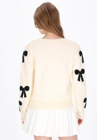Krämfärgad stickad tröja med svarta rosettmönster på ärmarna, texturerad med subtila glittern. Ribbad mudd och nederkant. I kombination med en veckad vit kjol.