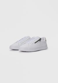 Hvite lave sneakers med hvite snøringer og glidelås på siden, med et minimalistisk design og subtil merkevaretekst på siden.