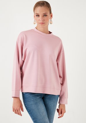 Sudadera rosa claro con cuello alto, mangas largas y textura suave. Combinada con jeans azules, que muestran un ajuste relajado y un diseño moderno.