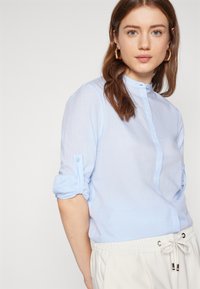 Camicia azzurro chiaro con colletto alla cinese, maniche arrotolate e una trama trasparente, abbinata a pantaloni chiari.